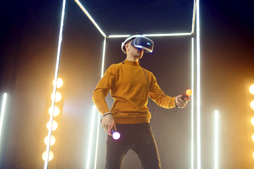 Male gamer plays the game using virtual reality headset and gamepad in luminous cube. Dark playing club interior, spotlight on background, VR technology with 3D vision. Male gamer using virtual reality headset
