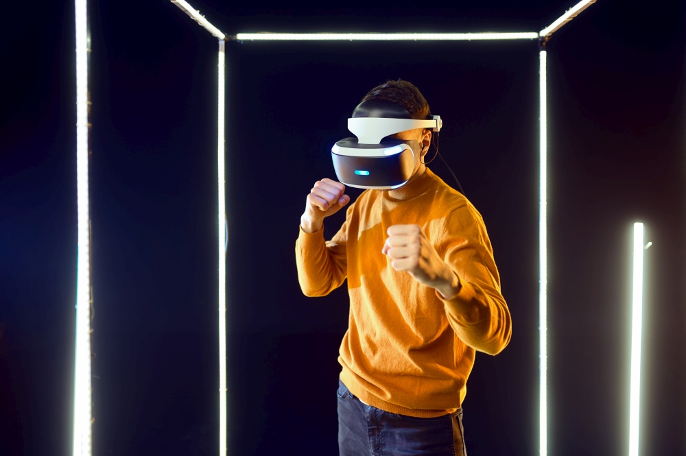 Young gamer fighting in virtual reality headset and gamepad in luminous cube, front view. Dark playing club interior, spotlight on background, VR technology with 3D vision. Young gamer fighting in virtual reality headset