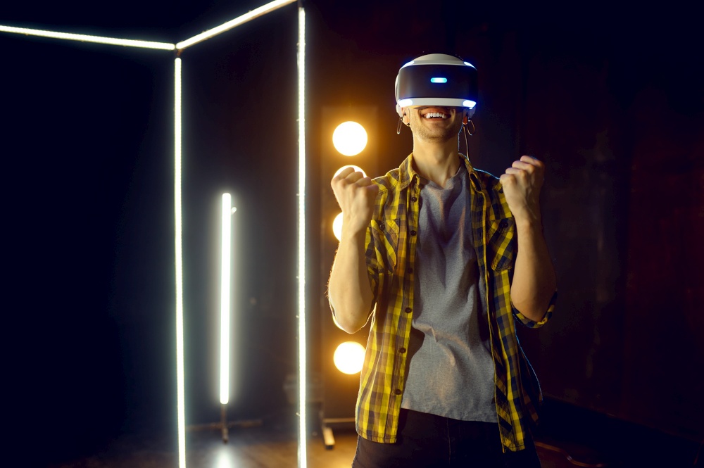 Happy man using virtual reality headset and gamepad in luminous cube, front view. Dark playing club interior, spotlight on background, VR technology with 3D vision. Happy man using virtual reality headset