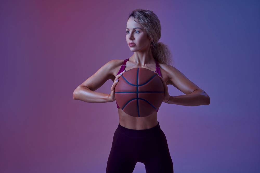 Young sportswoman with ball poses in studio, neon background. Fitness woman at the photo shoot, sport concept, active lifestyle motivation. Young sportswoman with ball poses in studio