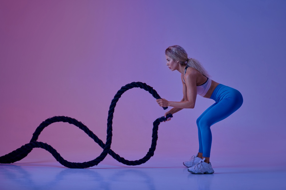 Young sportswoman with ropes poses in studio, neon background, crossfit. Fitness woman at the photo shoot, sport concept, active lifestyle motivation. Young sportswoman with ropes in studio, crossfit