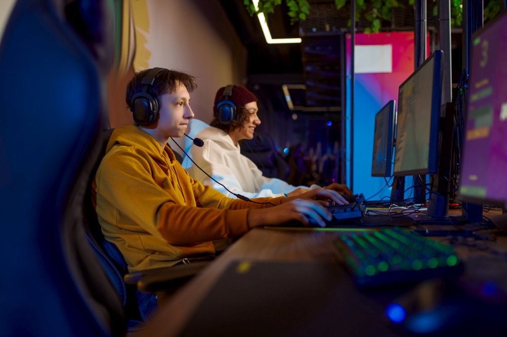 Two young gamers in headsets play in video game club. Virtual entertainment, e-sport tournament, cybersport lifestyle. Male person leisures in internet cafe. Two young gamers in headsets play in video game club