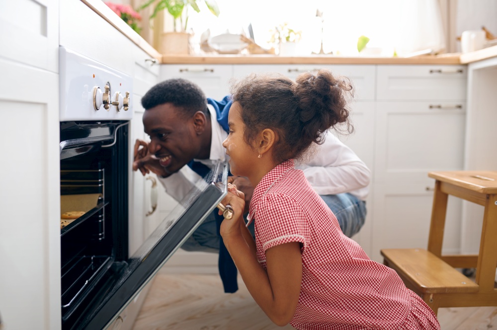 Happy father and little kid cooking cakes in oven on breakfast. Smiling family on the kitchen in the morning. Dad feeds female child, good relationship. Father and kid cooking cakes in oven on breakfast