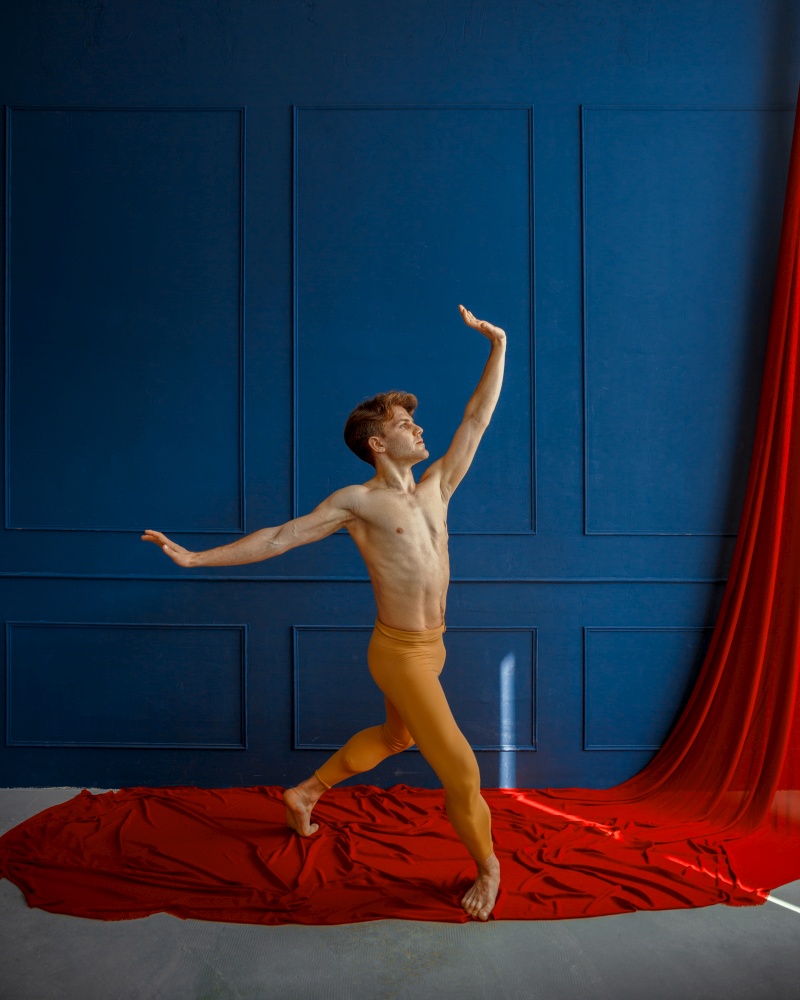 Male ballet dancer, performing in action, dancing studio, blue wall and red cloth on background. Performer with muscular body, grace and elegance of movements. Male ballet dancer, performing in action