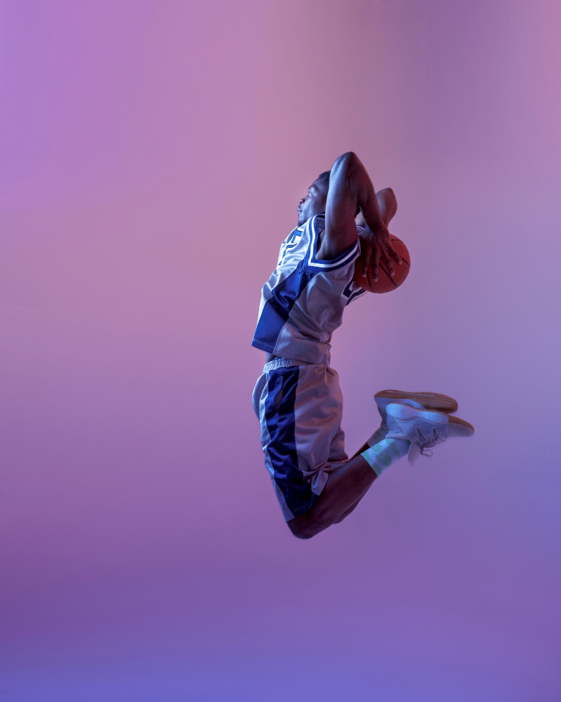 Basketball player jumping with ball in studio, neon background. Professional male baller in sportswear playing sport game. Basketball player jumping with ball in studio