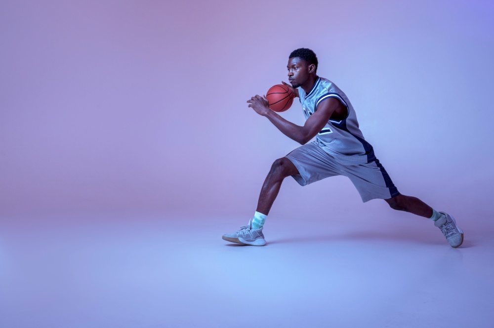 Basketball player practicing with ball in studio, neon background. Professional male baller in sportswear playing sport game, sportsman. Basketball player practicing with ball in studio
