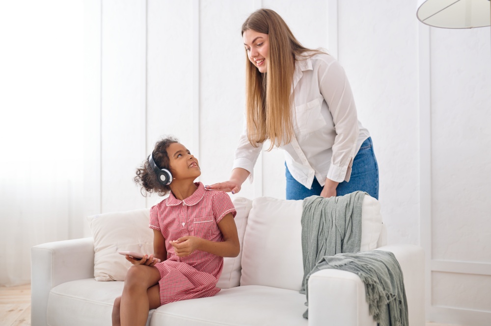 Mother and her little daughter in headphones on sofa in living room.Mom and female child leisures in their house together, good relationship, parental care. Mother and her little daughter in headphones