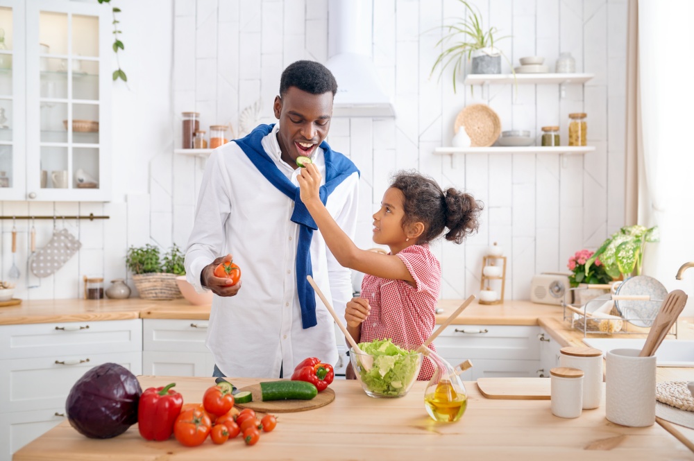 Happy dad and kid cooking vegetable salad on breakfast. Smiling family eats on the kitchen in the morning. Father feeds female child, good relationship. Dad and kid cooking vegetable salad on breakfast