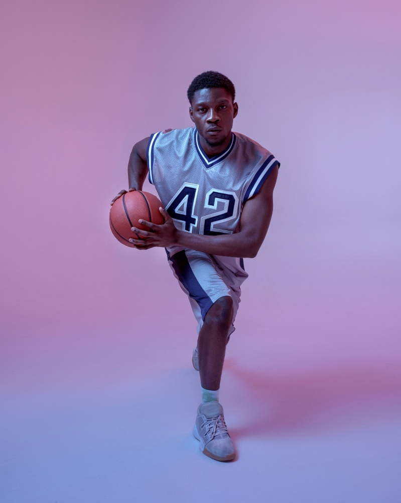 Basketball player practicing with ball in studio, neon background. Professional male baller in sportswear playing sport game.. Basketball player practicing with ball in studio