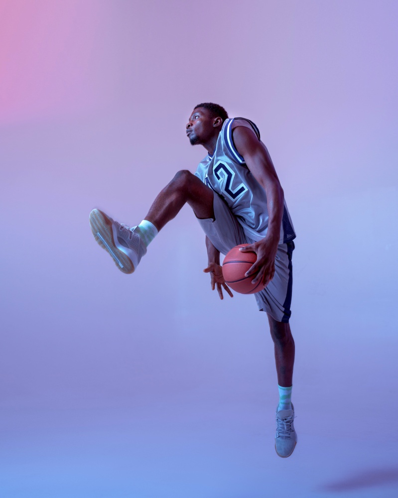 Basketball player moving with ball in studio, neon background. Professional male baller in sportswear playing sport game, tall sportsman. Basketball player moving with ball in studio