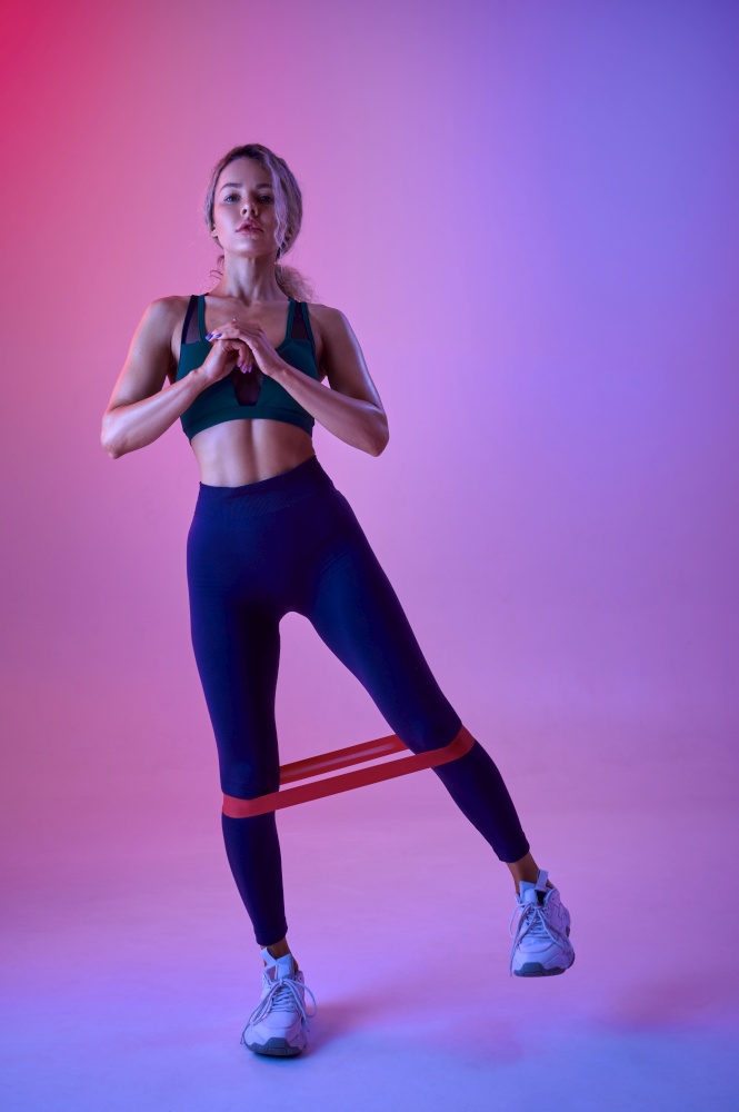 Athletic sportswoman doing exercise with rubber in studio, neon background. Fitness woman at the photo shoot, sport concept, active lifestyle motivation. Athletic sportswoman doing exercise with rubber