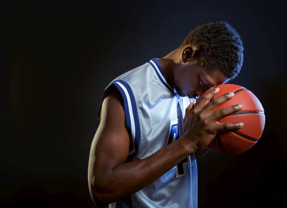 Focused basketball player poses with ball in studio, black background. Professional male baller in sportswear playing sport game, tall sportsman. Focused basketball player with ball in studio