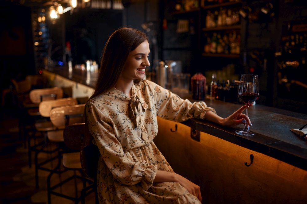 Alone young woman with glass of red wine sitting at the counter in bar. One female person in pub, human emotions, leisure activities, night life. Alone woman with glass of wine sitting in bar