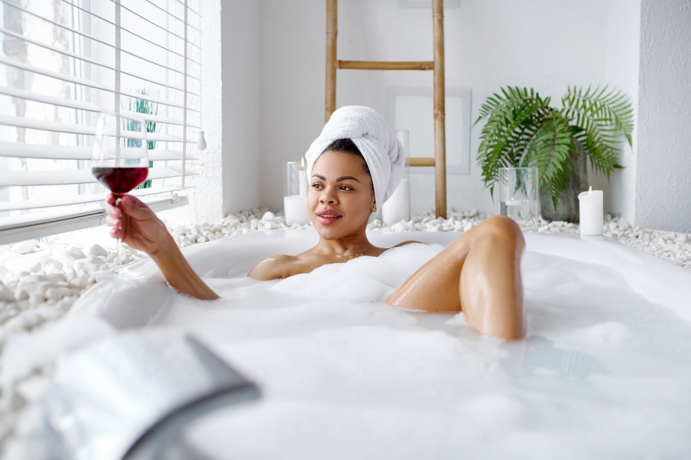 Seductive woman with glass of wine relax in a bubble bath. Female person in bathtub, beauty and health care in spa, wellness treathment in bathroom, pebbles and candles on background. Woman with glass of wine relax in a bubble bath