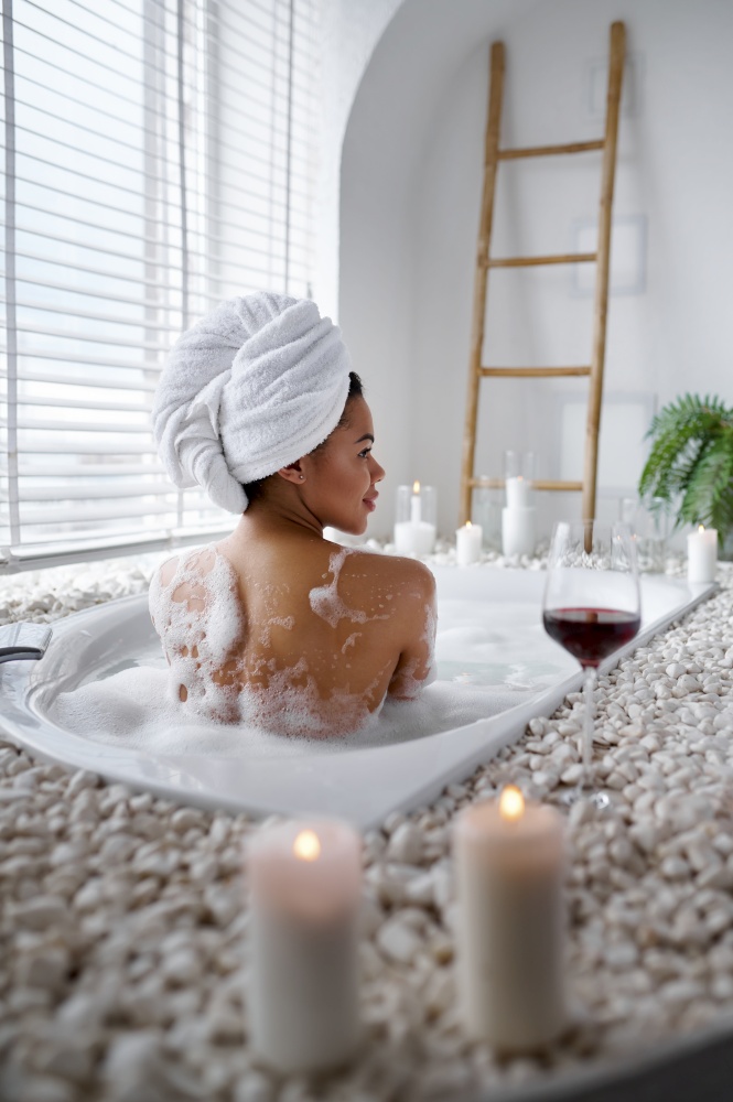 Seductive woman relax in a bubble bath, back view. Female person in bathtub, beauty and health care in spa, wellness treathment in bathroom, glass of wine, pebbles and candles on background. Seductive woman relax in a bubble bath, back view