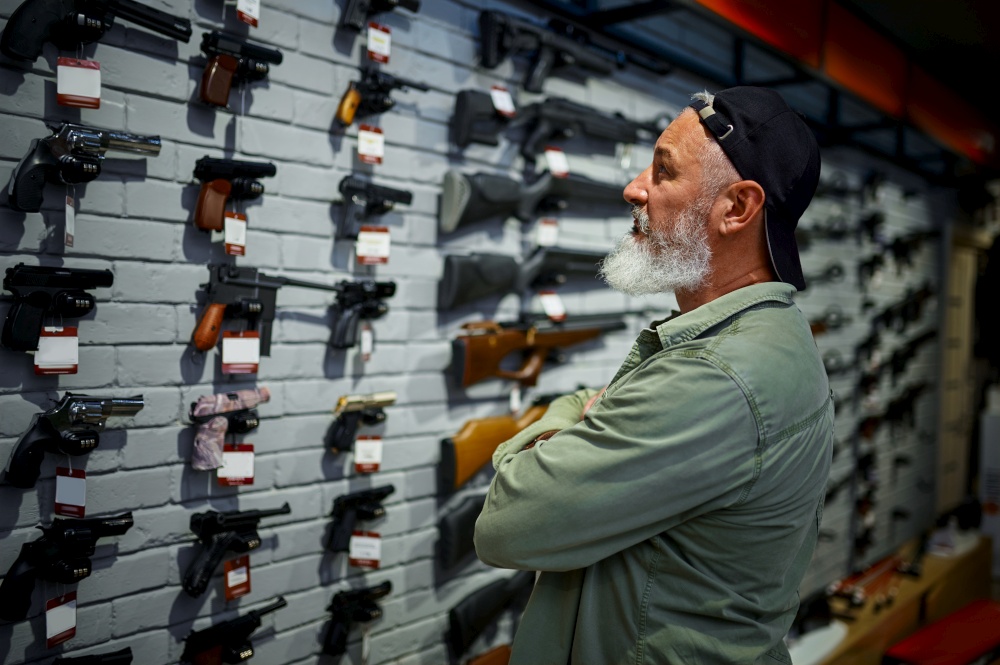 Bearded man choosing pistol at the showcase in gun store. Weapon shop interior, ammo and ammunition assortment, firearms choice, shooting hobby and lifestyle, self protection. Man choosing pistol at the showcase in gun store