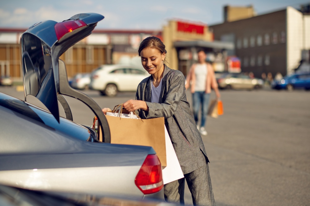 Woman puts her purchases in the trunk on supermarket car parking. Happy customer carrying purchases from the shopping center, vehicles on background. Woman puts her purchases in the trunk on parking