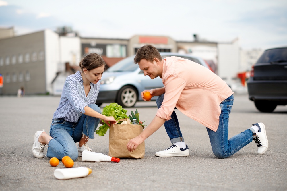 Family couple dropped the package with food on supermarket car parking. Happy customers carrying purchases from the shopping center, vehicles on background. Couple dropped the package on supermarket parking
