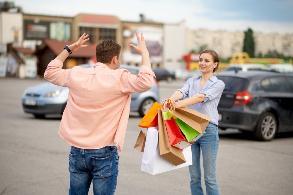 Husband is shocked by his wife&rsquo;s purchases, car parking. Happy customers with purchases near the shopping center, vehicles on background, family couple in market. Husband is shocked by wife&rsquo;s purchases, parking
