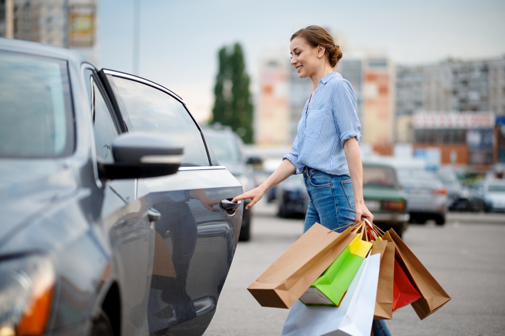 Woman puts her purchases in car on market parking. Happy customer carrying purchases from the shopping center, vehicles on background. Woman puts her purchases in car on market parking