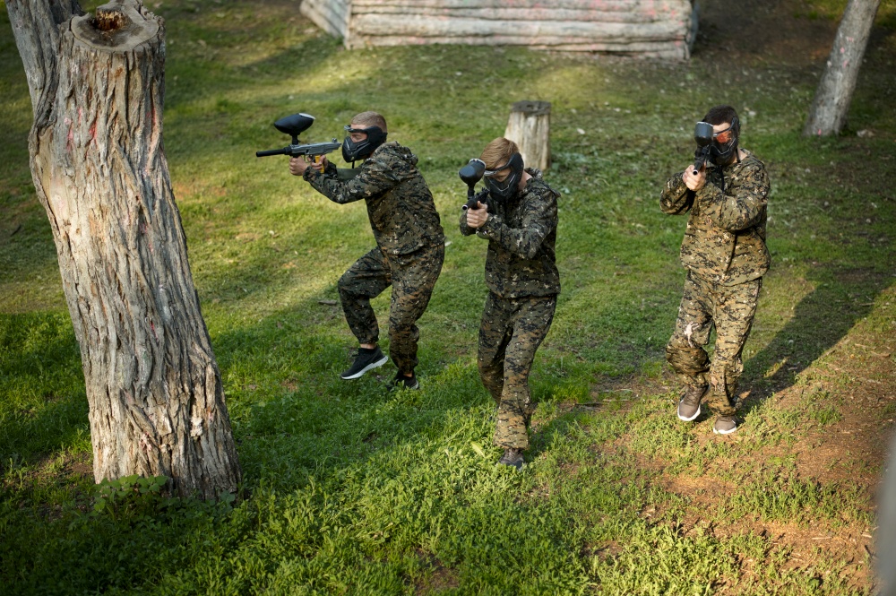 Team in uniform and masks playing paintball, battle on playground in the forest. Extreme sport with pneumatic weapon and paint bullets or markers, military game outdoors, combat tactics. Team in uniform playing paintball in the forest