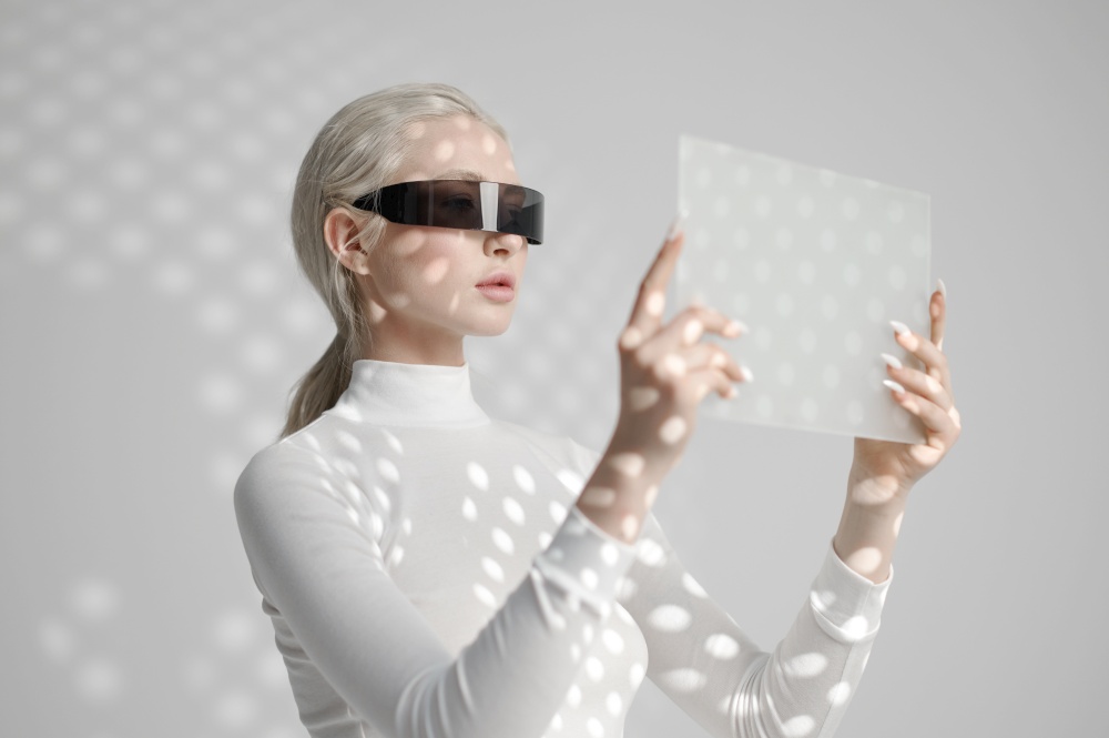 Young woman in white clothes and glasses holds futuristic gadget, light grey background. Female person in virtual reality style, future technology, futurism concept. Young woman in glasses holds futuristic gadget