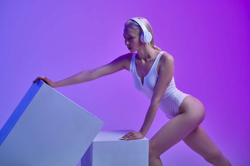 Sportive futuristic woman in white sportswear and modern headphones stands at the cubes, purple background. Sexy female person in virtual reality style, future technology, futurism concept. Sportive futuristic woman in modern headphones