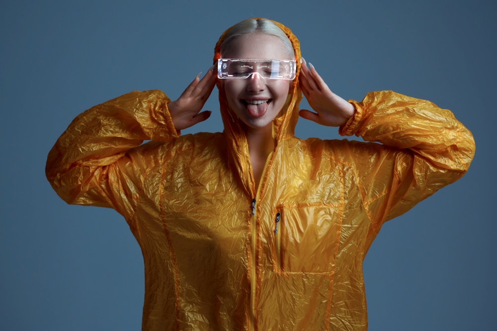 Funny futuristic woman in raincoat and modern glasses shows her tongue, grey background. Sexy female person in virtual reality style, future technology, futurism concept. Funny futuristic woman shows her tongue, futurism