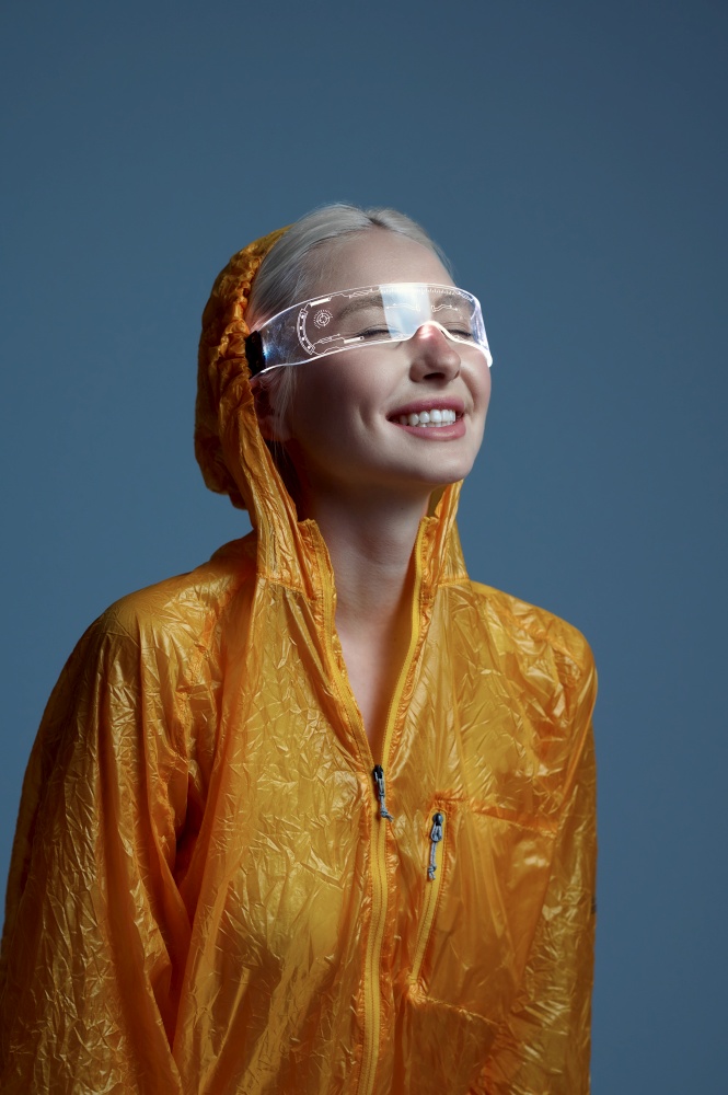 Futuristic young woman in raincoat and modern glasses, grey background. Sexy female person in virtual reality style, future technology, futurism concept. Futuristic woman in raincoat and modern glasses