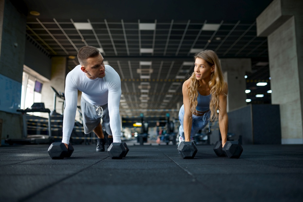 Sportive couple doing push-ups with dumbbells, training in gym. Athletic man and woman on workout in sport club, active healthy lifestyle. Couple doing push-ups with dumbbells in gym