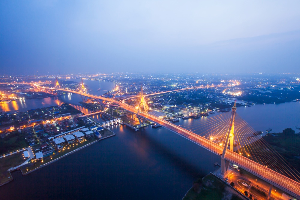 Aerial view of Bhumibol Suspension Bridges and highways interchange over the Chao Phraya River at dusk, beautiful light trails across the bridges and river curve. Samut Prakan, Thailand. Long exposure.