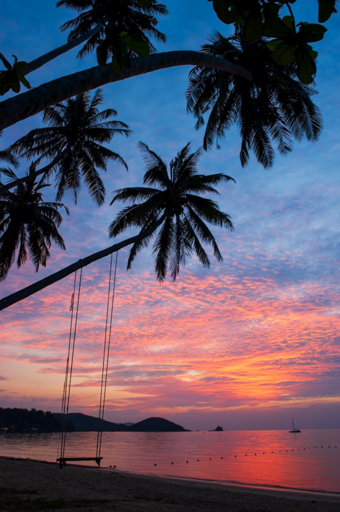 Fantastic clouds and sunset sky over the tropical island, beautiful shape of coconut palm trees and swing on the beach, serene scene on summer dusk. Koh Mak Island, Thailand.