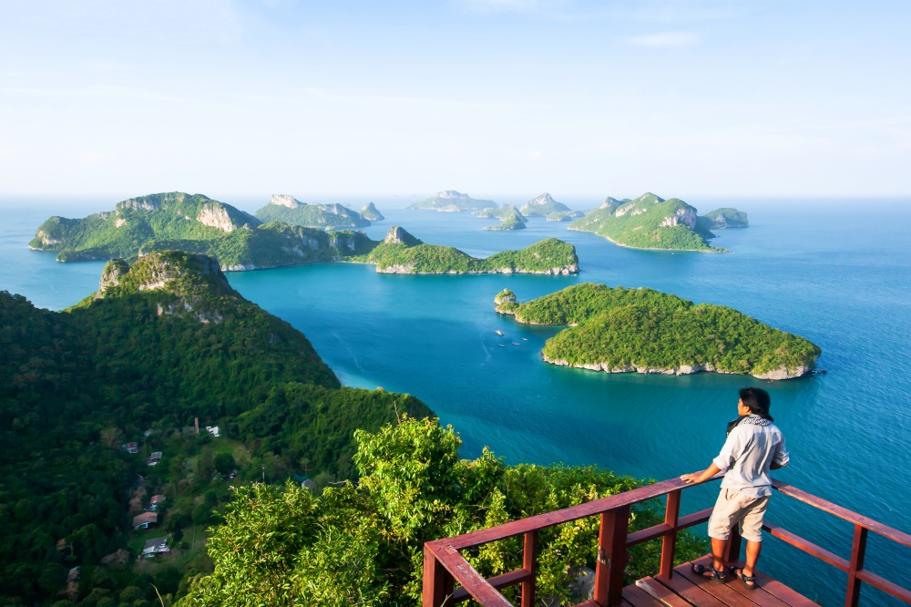 A young man in casual standing alone on viewpoint at the top of Koh Wua Ta Lap while enjoying fantastic scenery of blue sea with lots of tropical islands, picturesque on sunny summer.