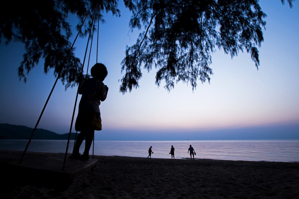A little local girl playing on the swing and looking at a group of tourists walking on the beach at sunset. Koh Chang Island, Ranong, Thailand.