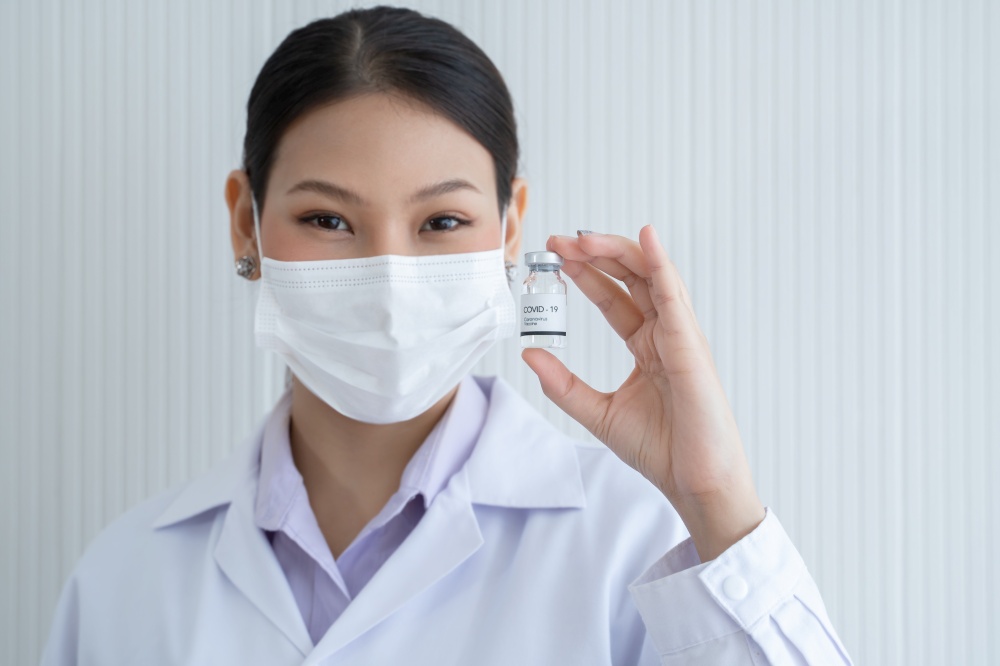 Selective focus on Covid-19 vaccines vial bottle in hand of Asian young doctor woman wear face mask, white gown and smiling on white background. Close up portrait
