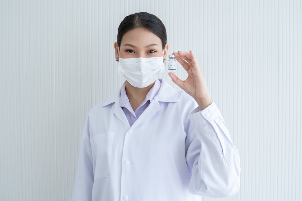 Portrait of Asian young doctor or scientist woman holding showing Covid-19 vaccines vial bottle in hand wear face mask, white gown and smiling. White background