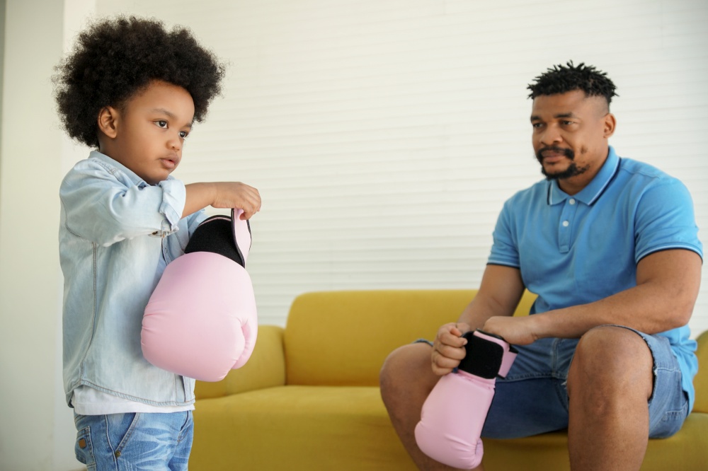 African American family father and little son playing boxing together. Young black Father sitting on sofa watching little boy wearing pink boxing gloves at home