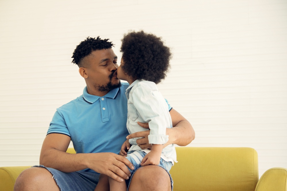 African American family little son kiss his young father at lip while sitting on sofa at home