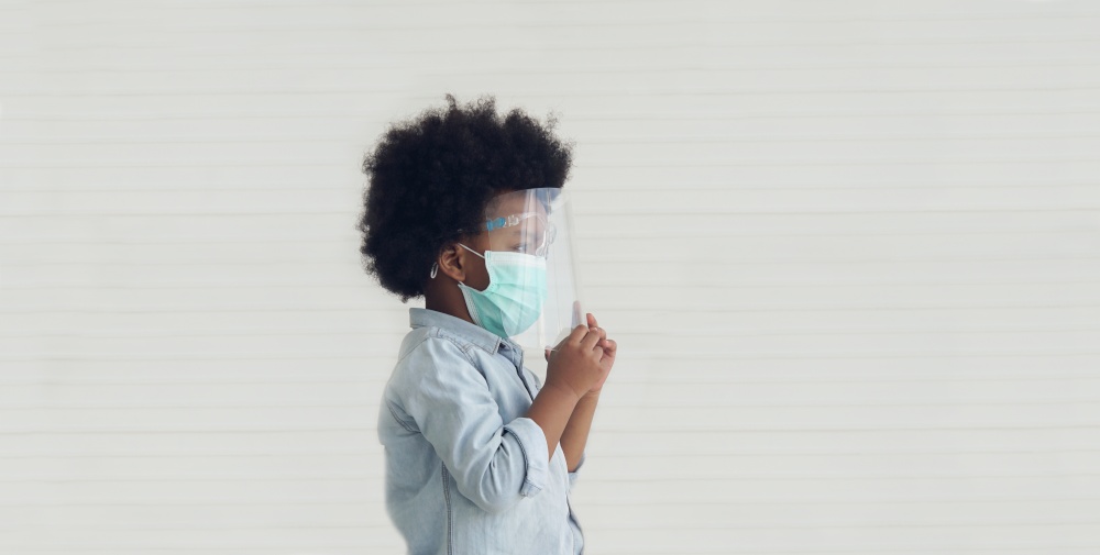 Side view of little African American boy wearing mask and face shield standing looking outside on white background