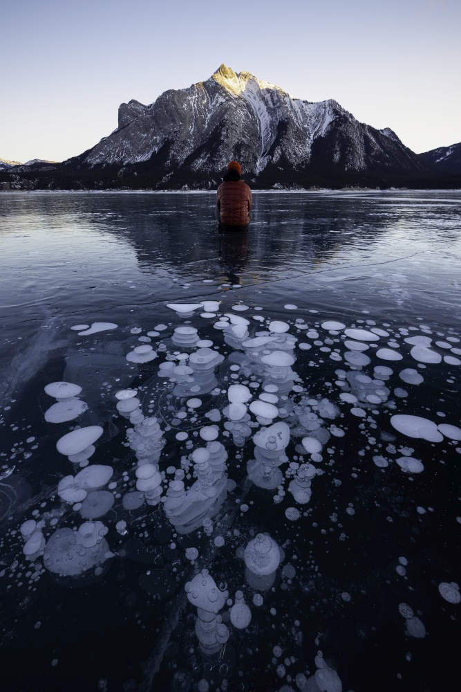 An adventurer sits in front of an abstract array of the frozen ice bubbles of Abraham Lake as the sun sets on the day.
