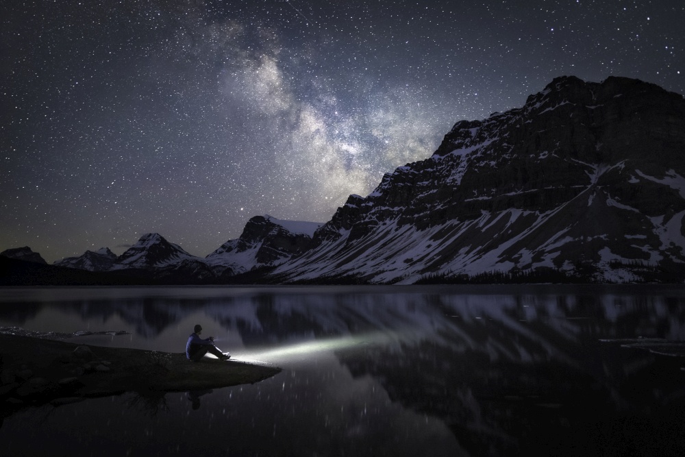 The Milky Way shining over Bow Lake and Crowfoot Mountain alongside the Icefields Parkway in Banff National Park while a stargazer illuminates the blue-green waters of the icy lake.