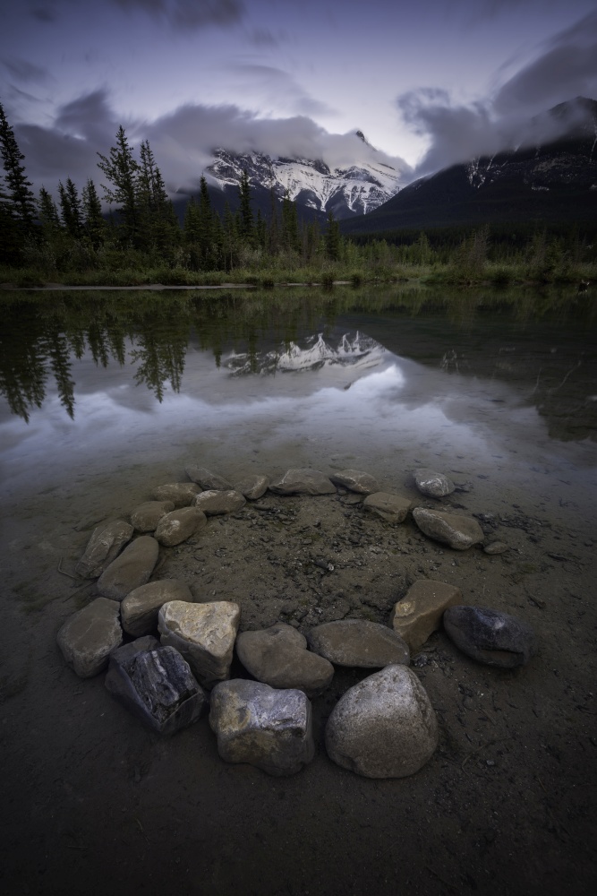 A ring of rocks submerged under a creek in Canmore as the Three Sisters reflect in the water below during a Summer evening.