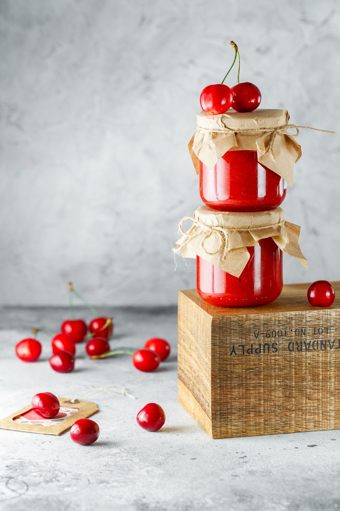 Homemade cherry jam in glass jar on the wooden box on the gray background. Two jars of cherry jam on wood with cherries in the back. Food photography. Seasonal cooking concept