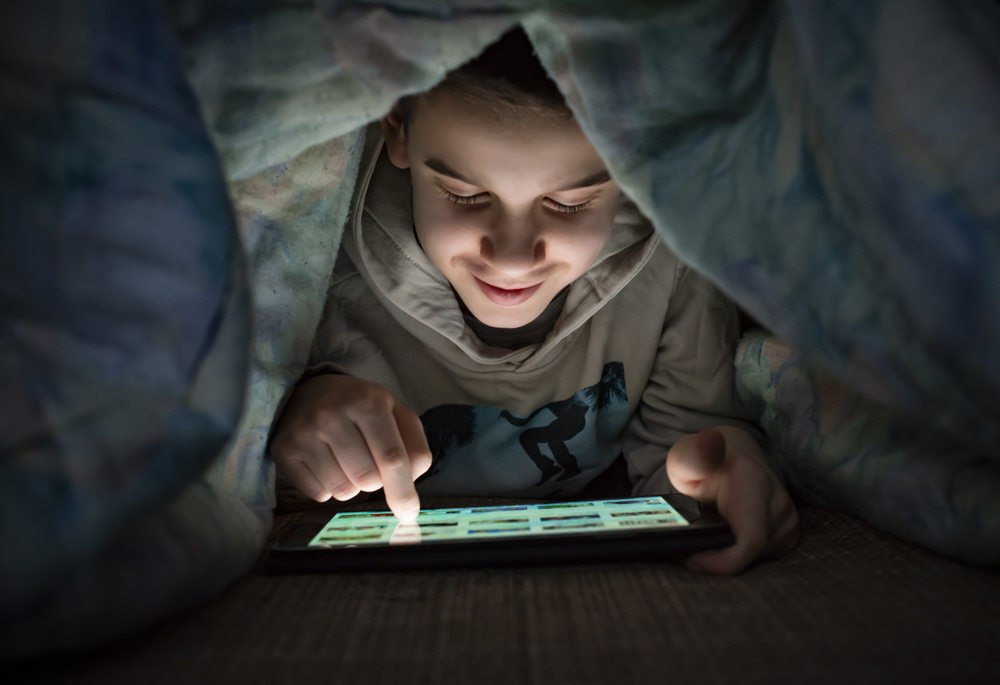 Child watching his tablet in the bed. Illuminated child face from device screen. Smiling Boy under the covers hold a tablet. Night time.