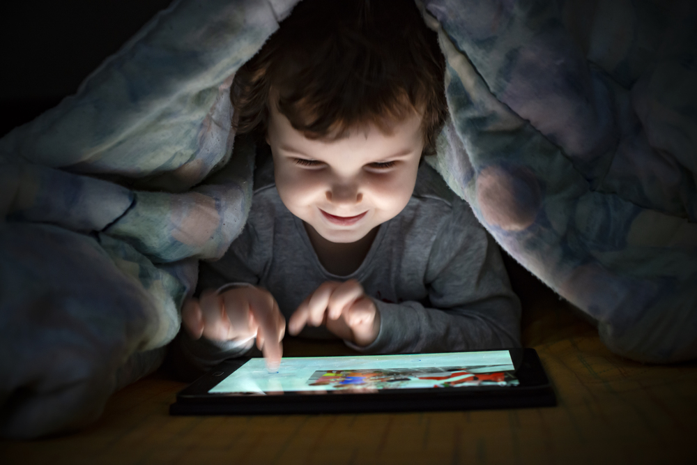 Little girl watching her tablet in the bed. Illuminated smiling child face from device screen. Child dressed with pajamas under the covers hold a tablet. Night time.