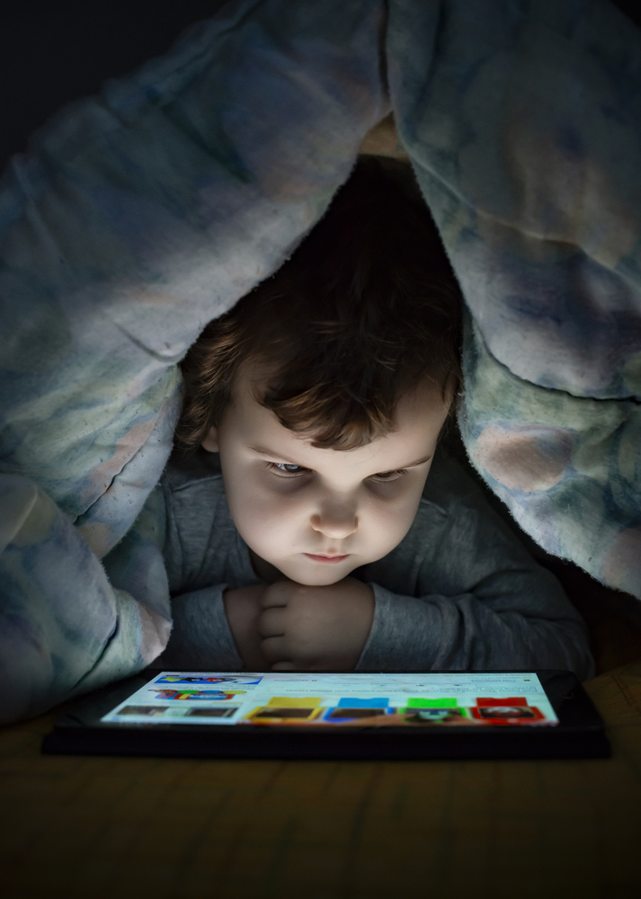 Little girl watching her tablet in the bed. Illuminated child face from device screen. Child dressed with pajamas under the covers hold a tablet. Night time.