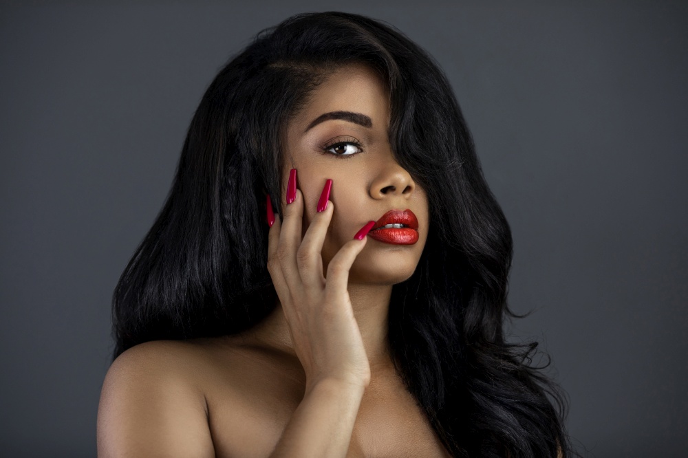 A portrait of a young Latina with long black wavy hair, beautiful makeup, red lips and nails posing by herself in a studio with a grey background.