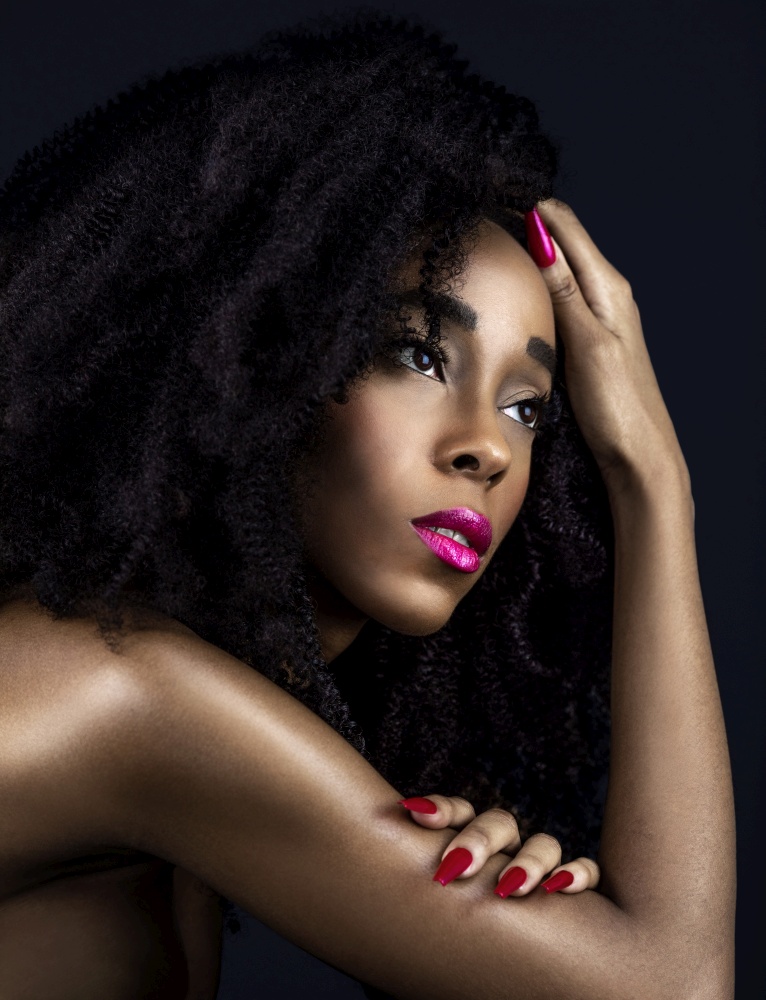 A portrait of a sensual young black female with long black curly hair, beautiful makeup, fuchsia lips and nails posing by herself in a studio with a dark background.