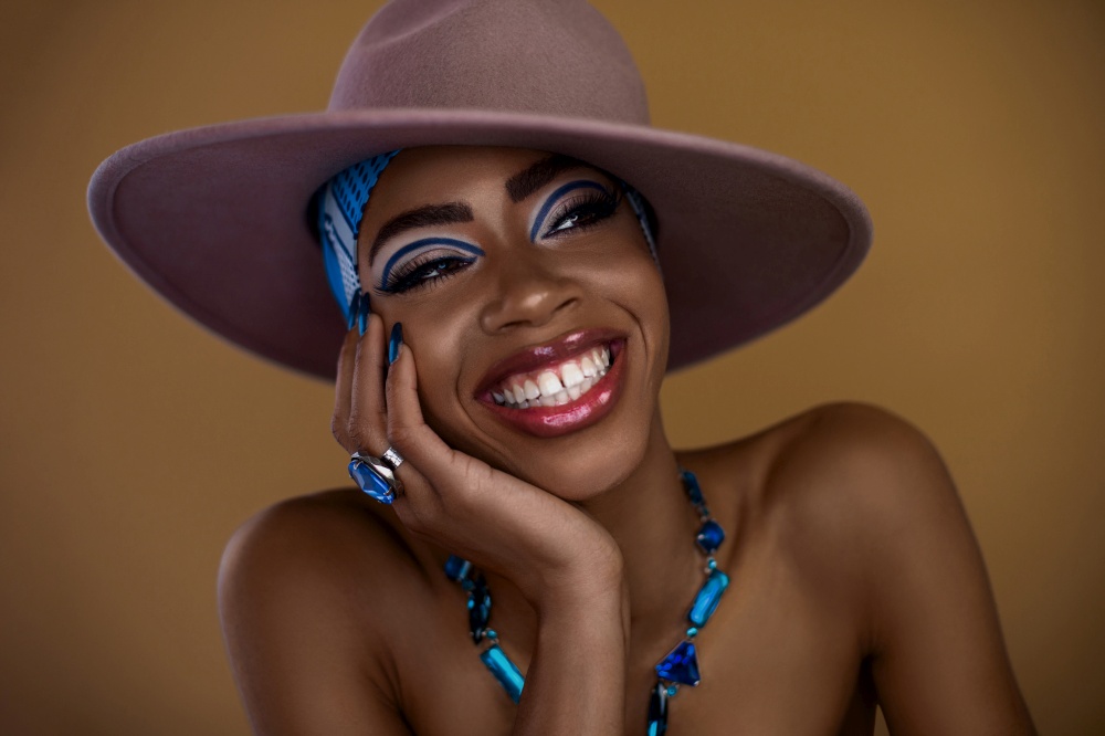 A beautiful young black female with a gorgeous smile & makeup posing by herself inside a studio wearing a grey hat, blue head scarf and jewelry.