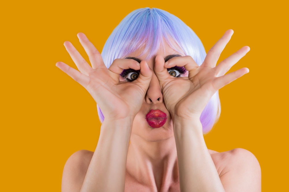 Young glam girl in wig and with bright makeup looking through fingers at camera grimacing. . Bright model making faces at camera